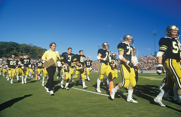 Photo shows a team of football players in uniform and their coach at a football stadium.