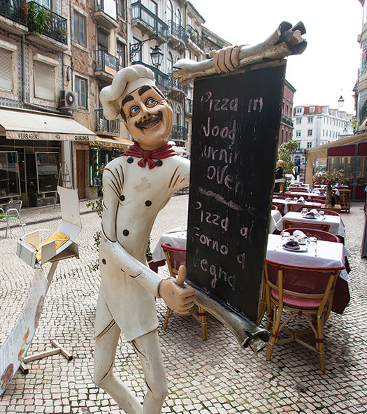 Photo shows a mannequin dressed like a chef, holding a menu card on the sidewalk in front of a restaurant.