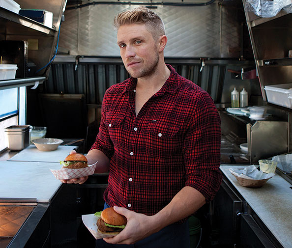 Photo shows a man holding a burger in ach of his hands inside a food truck.