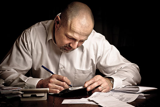 Photo shows a man seated at a desk and writing in a book. Financial statements are all around him.