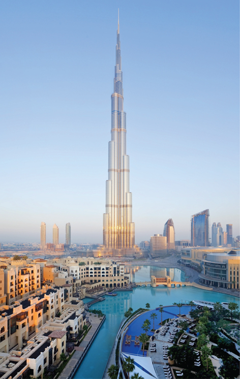 The Burj Khalifa building stands as a tall spire surrounded by shorter buildings. In the foreground are a river and some palm trees.