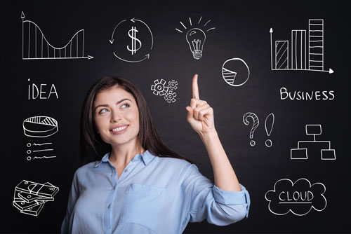 Photo shows a woman standing in front of a blackboard and smiling with her index finger raised. On the board are several images denoting a cloud, an idea, a bar graph, money, and so on.