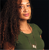 A photograph of a woman in studio light. She has curly hair and wears a thin necklace.