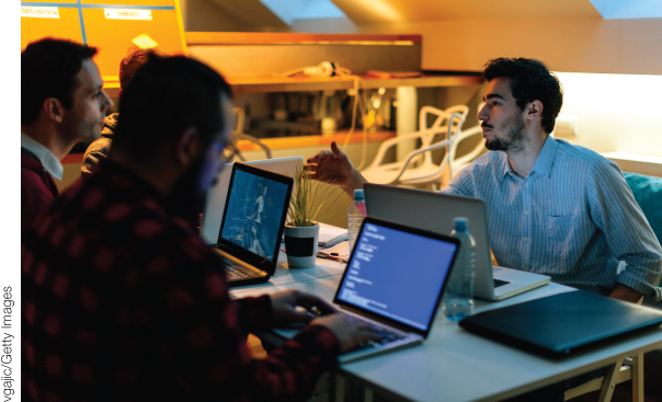 Photo illustration of three men working on their laptops. One of the them is discussing with another man seated on the opposite side.