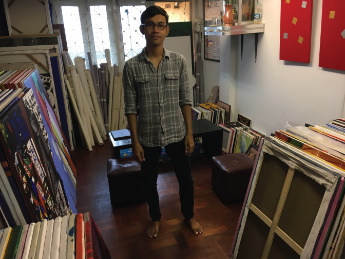 Photograph of a boy, a former monk, standing in his art gallery full of painting frames and rolls of canvas. 